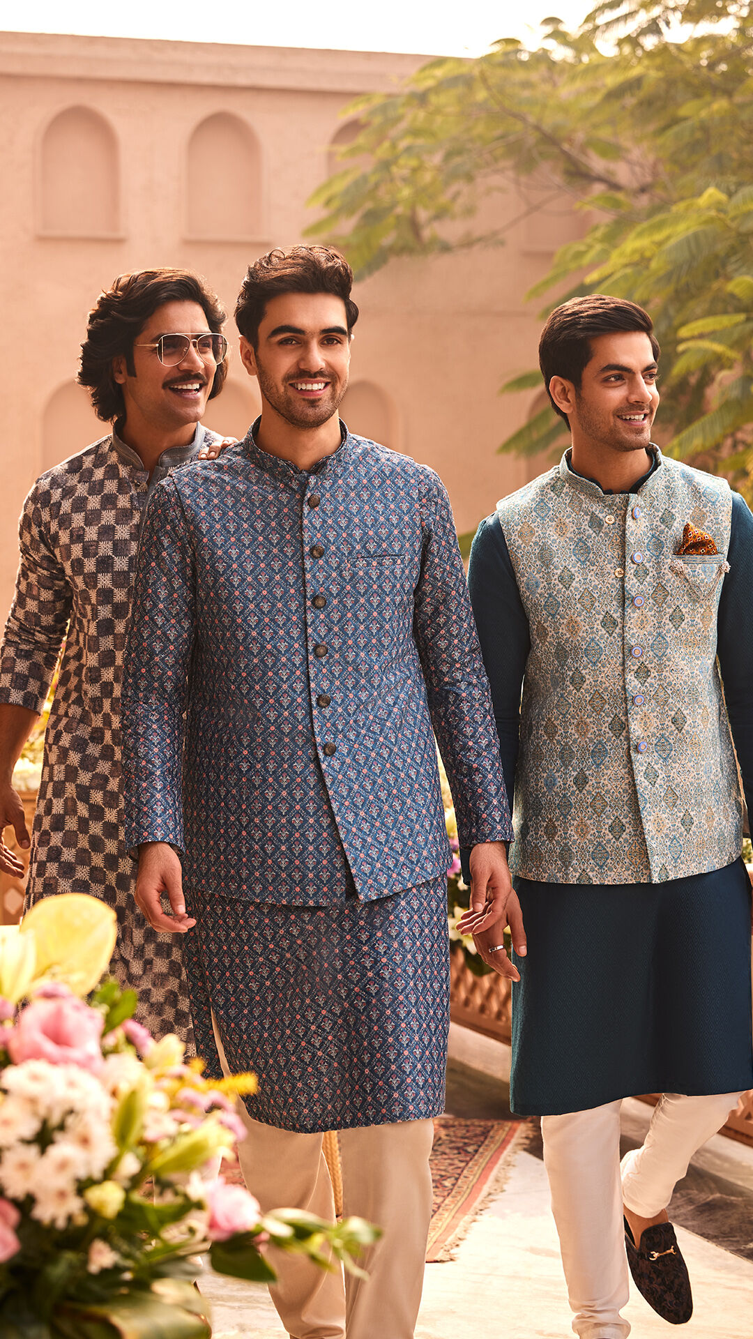 Three men in stylish traditional Indian ethnic wear, walking outdoors and smiling, featuring patterned kurtas in blue and grey tones.